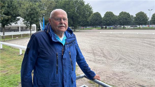 Ein Mann mit blauer Jacke steht an einem sandigen Reitplatz.
