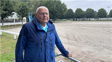 Ein Mann mit blauer Jacke steht an einem sandigen Reitplatz.