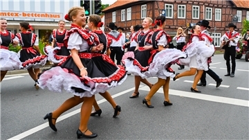 Schwungvoll tanzen mehrere Frauen in rot-weiß-schwarzer Tracht über die Straße.