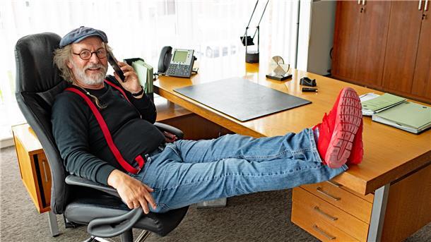 Ein Mann sitzt in einem Arbeitszimmer, telefoniert und hat die Füße auf dem Tisch liegen.