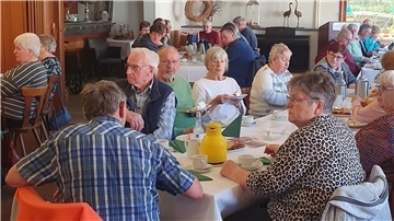 Überwiegend ältere Menschen sitzen an gedeckten Tischen, genießen Kaffee und Kuchen und scheinen einer Ansprache zu lauschen. 