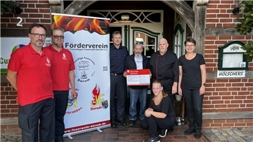 Eine Frau hält einen Scheck in der Hand. Drumherum stehen Männer der Feuerwehr.