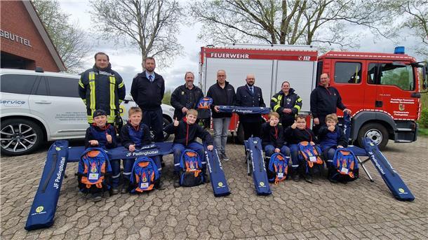 Vor einem Pkw und einem Einsatzfahrzeug der Feuerwehr Padingbüttel posieren sieben Erwachsene und sechs Kinder für ein Gruppenfoto mit neuen Liegen und Rucksäcken.