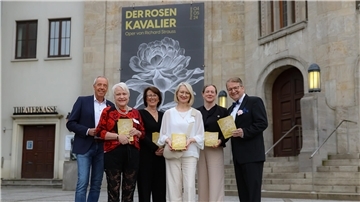 vier Frauen und zwei Männer stehen vor dem Eingang des Stadttheaters Bremerhaven, an dessen Fassade ein großes Plakat hängt mit der Aufschrift "Der Rosenkavalier – Oper von Richard Strauss"