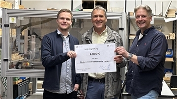 Drei Männer stehen in der Holzwerkstatt einer Schule und halten einen symbolischen Spendenscheck über 1000 Euro.