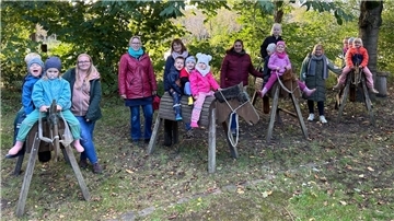 Eine Gruppe von Kindern sitzt auf hölzernen Spielpferden, die in einer Wiese unter Bäumen aufgestellt sind. Jedes Kind trägt warme Kleidung und Mützen, passend für kühleres Wetter. Hinter den Kindern stehen mehrere Erwachsene, die sie begleiten.