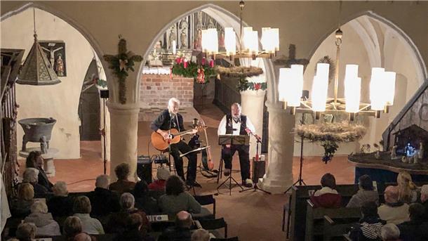 Ein stimmungsvoll beleuchtetes Konzert in einer historischen Kirche. Zwei Musiker spielen, einer mit Gitarre, der andere singend. Im Vordergrund sitzen Zuschauer in Kirchenbänken.
