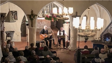 Ein stimmungsvoll beleuchtetes Konzert in einer historischen Kirche. Zwei Musiker spielen, einer mit Gitarre, der andere singend. Im Vordergrund sitzen Zuschauer in Kirchenbänken.