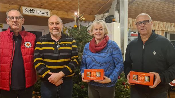 Vier Personen stehen nebeneinander vor einem geschmückten Weihnachtsbaum in einem Raum mit Holzdecke. Links trägt eine Person eine rote Jacke mit dem Logo des Deutschen Roten Kreuzes.