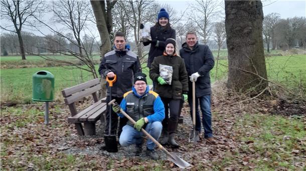 Eine Gruppe von fünf Personen steht und kniet in einem ländlichen Gebiet vor einer Sitzbank. Sie tragen warme Kleidung und Arbeitshandschuhe, passend für Garten- oder Landschaftsarbeiten. Einige halten Schaufeln und Gartengeräte in der Hand.