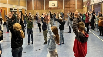 Eine große Gruppe von Kindern und Jugendlichen steht in einer Turnhalle und führt gemeinsam eine Übung aus. Alle haben die Arme nach oben gestreckt, während sie in Reihen aufgestellt sind.