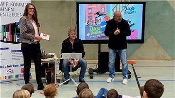 Drei Personen stehen und sitzen in einer Turnhalle vor einem Publikum von Kindern. Auf einem Monitor ist das Buchcover "Dachs und Rakete" zu sehen.