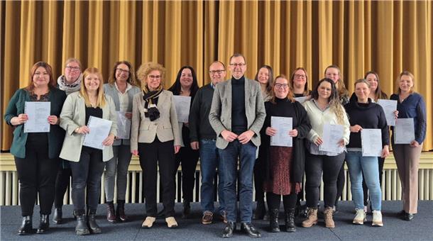 Eine Gruppe von 17 Personen, überwiegend Frauen, posiert auf einer Bühne vor einem goldfarbenen Vorhang. Viele der Personen halten Zertifikate oder Dokumente in der Hand und lächeln in die Kamera. Die Gruppe ist in gepflegter, teils geschäftlicher Kleidung gekleidet. 