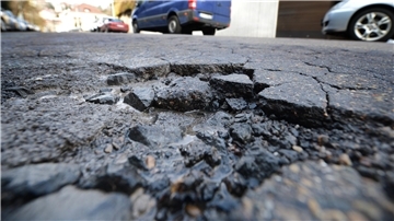 Schlagloch in einer Straße mit parkenden Autos im Hintergrund.