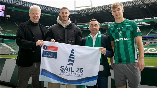 Das Foto ist im Weserstadion entstanden, es zeigt vier Herren, die eine Flagge in der Hand halten, die auf die Sail 2025 hinweist. Freuen sich über die Partnerschaft: Dr. Ralf Meyer vom SAiL-Organisationskomitee, die Spieler Marco Friedl und Keke Topp sowie Michael Gerber, Geschäftsführer der Erlebnis Bremerhaven GmbH.