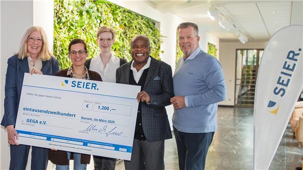 Fünf Personen stehen in einem modernen Innenraum mit begrünten Wänden und halten gemeinsam einen großen Spendenscheck der Firma SEIER über 1.200 Euro für den Verein GEGA e.V.