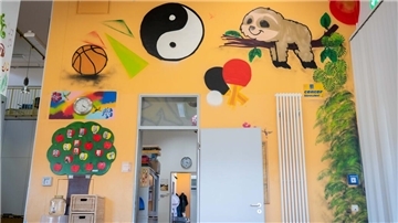 Eine farbenfrohe, kreativ gestaltete Wand in einem Innenraum. Die Wand ist gelb-orange gestrichen und mit verschiedenen Graffiti verziert, darunter ein Yin-Yang-Symbol, ein Faultier auf einem Ast, ein Basketball, Tischtennisschläger und grüne Pflanzen.