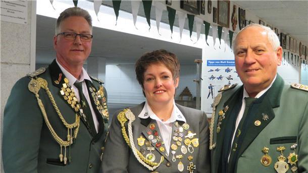 Drei Mitglieder eines Schützenvereins in festlicher Uniform mit zahlreichen Orden und Abzeichen stehen nebeneinander und lächeln in die Kamera. Im Hintergrund sind grün-weiße Wimpelketten und Schießstand-Zubehör zu sehen.