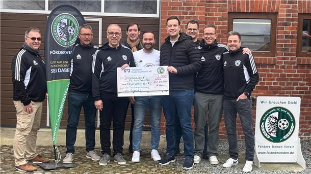 Eine Gruppe von Männern steht lächelnd vor einem Gebäude aus rotem Klinkerstein. Sie halten gemeinsam einen großen symbolischen Spendenscheck über 3.000 Euro, ausgestellt vom Förderverein des FC Land Wursten. Mehrere Männer tragen Vereinskleidung mit Vereinslogo. 