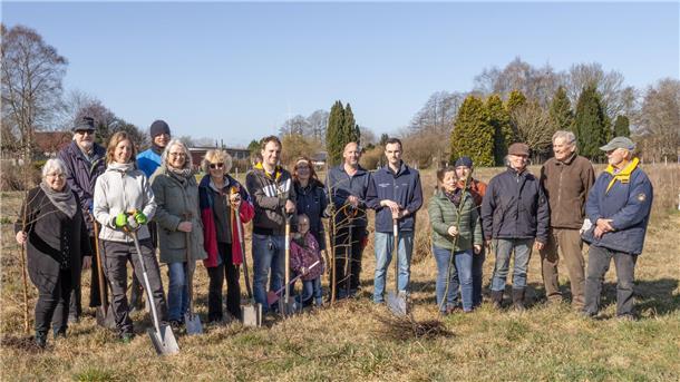 Eine Gruppe von 16 Menschen – von Kindern bis Senioren – steht auf einer Wiese an einem sonnigen Tag. Sie halten Schaufeln und junge Bäume.