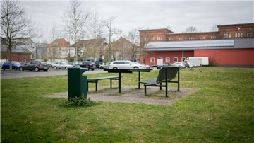 Im Vordergrund Rasen sowie zwei Bänke und ein Tisch, im Hintergrund ein mit Pkw belegter Parkplatz