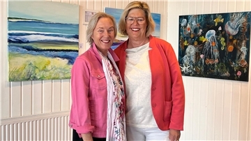 **ALT-Text:**  
Die Künstlerinnen Antje Boedecker und Marina Zander stehen lächelnd nebeneinander in einer Ausstellung vor ihren farbenfrohen Gemälden im Atrium Burhave.