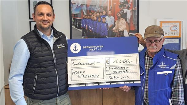 Zwei Männer stehen nebeneinander und halten gemeinsam einen überdimensionalen blauen Scheck über 1.000 Euro zugunsten von „Bremerhaven Hilft e.V.“, im Hintergrund hängt ein Bild von Jugendfußballern.