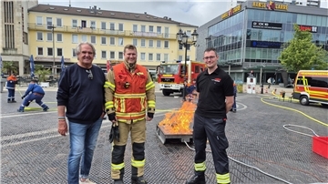 Drei Männer stehen nebeneinander. Im Hintergrund sind Flammen, ein Feuerwehrfahrzeug und ein Rettungswagen zu sehen.   