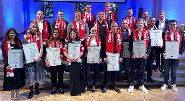 Gruppenfoto von rund 20 frischgebackenen Handwerksmeistern und -meisterinnen bei der Meisterfeier in Bremen. Die Personen stehen auf einer Bühne vor blau beleuchtetem Hintergrund und tragen rote Schals mit der Aufschrift „DAS HANDWERK – DIE WIRTSCHAFTSMACHT VON NEBENAN.“ Alle halten stolz ihre Meisterbriefe in den Händen. 