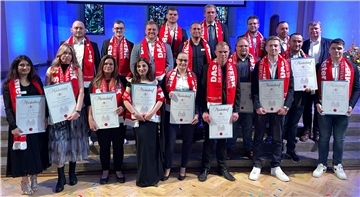 Gruppenfoto von rund 20 frischgebackenen Handwerksmeistern und -meisterinnen bei der Meisterfeier in Bremen. Die Personen stehen auf einer Bühne vor blau beleuchtetem Hintergrund und tragen rote Schals mit der Aufschrift „DAS HANDWERK – DIE WIRTSCHAFTSMACHT VON NEBENAN.“ Alle halten stolz ihre Meisterbriefe in den Händen. 