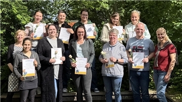 **ALT-Text:**
Zehn frisch zertifizierte Kindertagespflegepersonen stehen mit ihren Zertifikaten und je einer Rose in der Hand in zwei Reihen vor grünem Hintergrund und lächeln in die Kamera. Links und rechts stehen zwei Vertreterinnen der VHS.