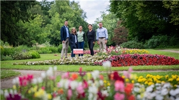 Fünf Personen stehen in einem bunt bepflanzten Blumenbeet im Bürgerpark. Im Vordergrund und Hintergrund blühen zahlreiche Sommerblumen in kräftigen Farben. Ein Schild weist auf eine Spende der Weser-Elbe Sparkasse hin. Die Szene ist von grünem Parkgelände und Bäumen umgeben.
