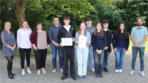 Gruppenfoto im Freien: Schüler:innen und Lehrkräfte der BBS Cuxhaven stehen gemeinsam, zwei halten Urkunden und eine Auszeichnung als „Nachhaltige Schülerfirma“.