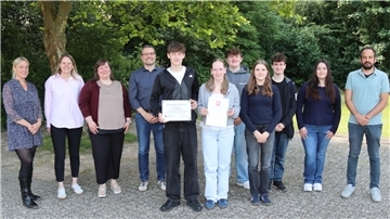 Gruppenfoto im Freien: Schüler:innen und Lehrkräfte der BBS Cuxhaven stehen gemeinsam, zwei halten Urkunden und eine Auszeichnung als „Nachhaltige Schülerfirma“.