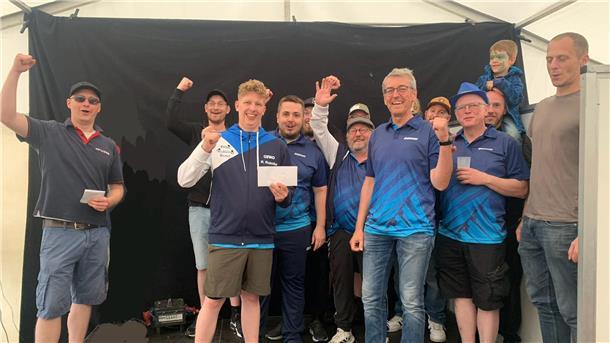 Gruppe jubelnder Männer, teils in blauen Sporttrikots, steht vor schwarzem Hintergrund in einem Festzelt. Zwei Personen halten Umschläge, ein Kind mit Gesichtsbemalung sitzt auf den Schultern.