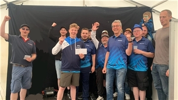 Gruppe jubelnder Männer, teils in blauen Sporttrikots, steht vor schwarzem Hintergrund in einem Festzelt. Zwei Personen halten Umschläge, ein Kind mit Gesichtsbemalung sitzt auf den Schultern.