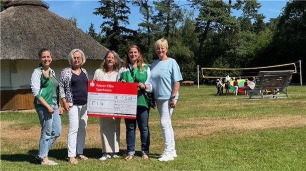 Fünf Frauen stehen auf einer Wiese vor einem runden Reetdachhaus und halten gemeinsam einen großen symbolischen Spendenscheck der Weser-Elbe Sparkasse über 1500 Euro. Im Hintergrund spielen Kinder mit einem bunten Schwungtuch auf einem Volleyballfeld.