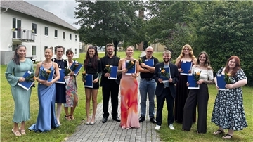 Zwölf frisch examinierte Pflegefachkräfte stehen mit Zeugnismappen und Blumen in der Hand lächelnd nebeneinander auf einem Weg im Grünen vor dem AMEOS Klinikum.