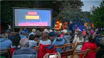 Menschen sitzen auf Stühlen vor einer Leinwand im Freien.
