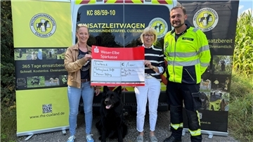 Drei Personen der Rettungshundestaffel Cuxland stehen vor einem Einsatzfahrzeug und halten einen symbolischen Spendenscheck über 1.000 Euro. Zwischen ihnen sitzt ein schwarzer Rettungshund. Im Hintergrund sind zwei Aufsteller mit Logos und Bildern der Staffel zu sehen.