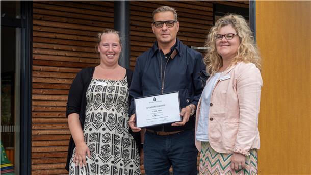 Drei Personen stehen lächelnd vor einem modernen Holzgebäude. Die mittlere Person hält eine gerahmte Spendenurkunde über 1.500 Euro der Proske-Stiftung in den Händen.