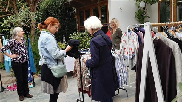 Mehrere Frauen stöbern an Kleiderständern im Freien nach Kleidung. Verschiedene Jacken, Blusen und Hemden hängen zum Tausch oder Kauf bereit.