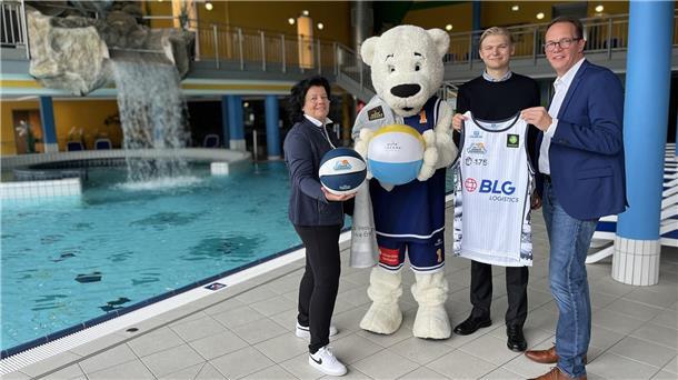 Drei Personen und ein Maskottchen in Eisbärenkostüm stehen am Beckenrand der Moor-Therme. Sie halten Basketbälle und ein Trikot, im Hintergrund ist ein Wasserfall zu sehen.