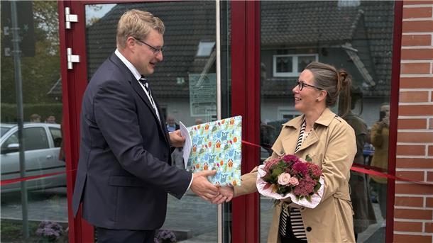 Schiffdorf weiht neue Kindertagesstätte IV ein Ein Mann im Anzug überreicht einer Frau mit Blumenstrauß vor einem Gebäude ein Geschenk. Beide lächeln, im Hintergrund ist ein rotes Band zur Eröffnung zu sehen.