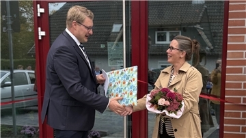 Ein Mann im Anzug überreicht einer Frau mit Blumenstrauß vor einem Gebäude ein Geschenk. Beide lächeln, im Hintergrund ist ein rotes Band zur Eröffnung zu sehen.