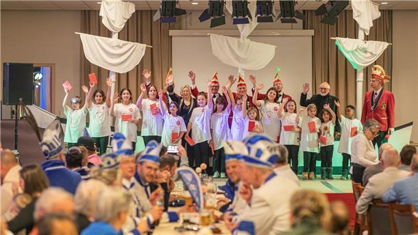 Gruppe von Kindern in weißen Shorts winken zusammen mit erwachsenen Karnevalisten mit Narrenkappen.