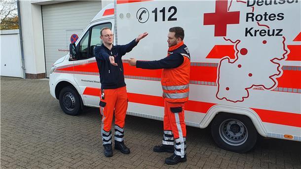 Zwei Sanitäter vor einem Rettungswagen, einer von ihnen hebt die Arme. 