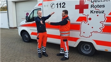 Zwei Sanitäter vor einem Rettungswagen, einer von ihnen hebt die Arme. 