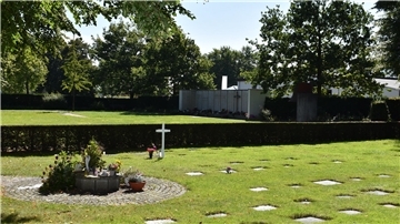 Blick auf eines der Rasenfelder auf dem Atenser Friedhof mit einer Blumen-Ablagefläche. Im Hintergrund ist das anonyme Gräberfeld mit Gedenkmauer zu sehen.