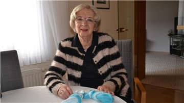 Eine grauhaarige Frau mit einer Brille hat vor sich gestrickte Mützen auf dem Tisch liegen.